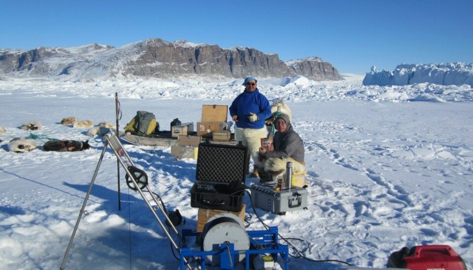 Hunters Lars and Rasmus take a break next to a system set up by Stefan Olsen from the Danish Meteorological Institute to measure ocean properties. The equipment is lowered into the sea, beneath the sea ice using a winch (foreground). The measurements will help them understand how sea ice is changing in the region. (Photo: Steffen Olsen/Francois Guerraz)