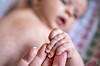 Nærbilde av babyhånd som holder en voksen finger, med et spedbarn i uskarp bakgrunn.