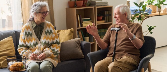 An older couple sit in a living room and chat while holding drinks beside a coffee table.