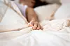 A hand grips a white bedsheet. The upper body of a woman can be seen. She has dark hair and is wearing a white top. The image is meant to depict her having an orgasm.