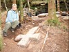 Irene Baug sitter ved siden av et stort steinkors som ligger på bakken. I bakgrunnen ses trær og avfallshaugen til det gamle steinkvernsbruddet.