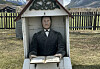 A small, chapel-shaped headstone featuring a sculpture and an open book, situated on a lawn in front of mountains and a fence.