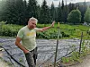 Farmer Erik Røed gestures beside vegetable beds in a forested rural garden.
